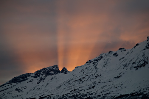 Bilde av solstråler som kommer opp bak snødekt fjell i Nord Norge